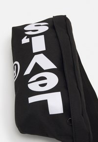 Svart tygväska med vit text och logotyp, rektangulär form, dragkedja och axelrem. Slitstarkt material med strukturerad yta.