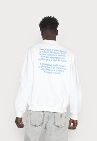 Felpa bianca con testo blu sul retro, scollatura rotonda e polsini a costine. Il testo esprime messaggi riguardanti l'indipendenza e i sogni.