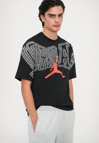 Μαύρη μπλούζα με κοντά μανίκια και έντονο άσπρο κείμενο "JORDAN", περιλαμβάνει μια κόκκινη σιλουέτα καλαθοσφαιριστή. Στρογγυλή λαιμόκοψη, άνετη εφαρμογή.