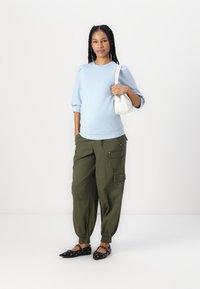 Suéter ajustado azul claro com mangas bufantes; calças cargo verdes oliva com bolsos; sapatilhas pretas com detalhes decorativos; pequena mala branca.