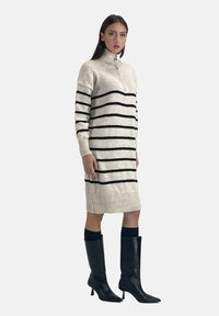 Strickkleid in Creme mit schwarzen horizontalen Streifen, hohem Kragen mit Reißverschluss, gerippten Bündchen und Saum. Kombiniert mit schwarzen kniehohen Stiefeln.