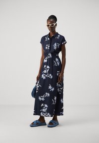 Abito maxi floreale navy con maniche a cappuccio, vita elastica e motivo floreale blu. Abbinato a sandali blu a scivolo e a una borsa blu lavorata.