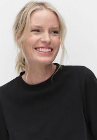 Musta, lyhythihainen paita, jossa on pyöreä pääntie, jota käyttää hymyilevä henkilö, jolla on löysästi aaltoileva vaalea hius, tasasävyistä taustaa vasten.