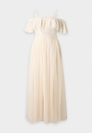 Femme portant une robe crème épaules dénudées avec un corsage à volants et une jupe transparente à pois, debout avec une expression neutre et des talons dorés.