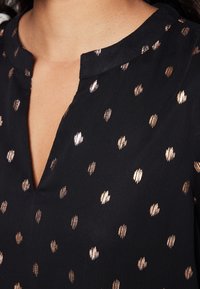 Blouse en tissu noir avec un col en V et un motif ponctué métallisé couleur or rose sur toute la surface. La texture semble lisse et légère.