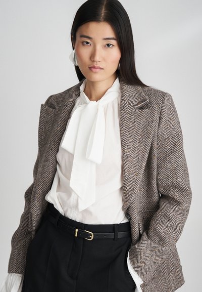 Houndstooth-kuvioinen bleiseri ruskeassa ja kermassa, valkoisella solmioblousella, mustilla housuilla ja kultaisella soljella varustettu vyö.