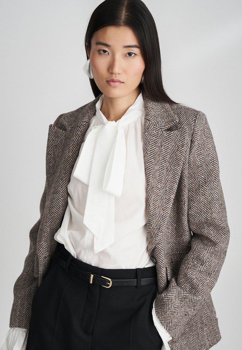 Blazer in pattern pied de poule marrone e crema, con una camicetta bianca con cravatta a farfalla, pantaloni neri e una cintura con fibbia dorata.