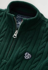 Suéter verde tejido con un cuello alto, que presenta una cremallera plateada y un patrón de punto de cable. Logo bordado en el pecho.