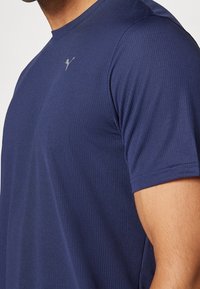 Navy modrá sportovní tričko s žebrovanou texturou, krátkými rukávy a malým logem na hrudi, určené pro aktivní nošení.