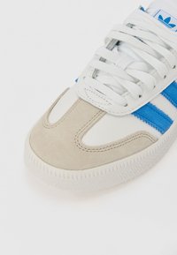 White sneaker with blue stripes, beige suede toe cap, white laces, and textured white rubber sole on a plain background.