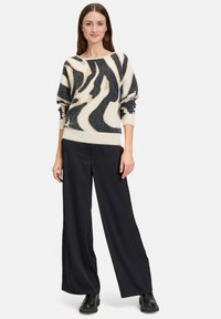Crème en zwarte oversized trui met abstract patroon, geribbelde manchetten en zoom; gecombineerd met wijde zwarte broek en flinke zwarte laarzen.