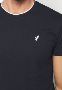 Marinblå bomullströja med rund vitt krage och en liten vit fågel broderad på vänster bröstområde. Slät textur, odekorerad design.