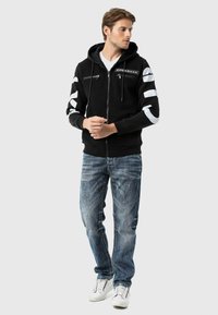 Schwarze Zip-up-Hoodie mit weißen Akzentstreifen an den Ärmeln, ausgestattet mit einer Vordertasche und einer Kapuze mit Zugband, kombiniert mit blauen Jeans und weißen Sneakern.