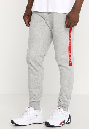 Ein Mann trägt hellgraue Jogginghosen mit rotem vertikalem "HILFIGER"-Schriftzug am linken Bein und weiße Sportschuhe mit schwarzen Sohlen.