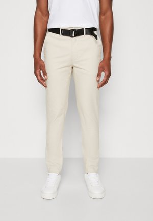 Pantaloni slim-fit beige chiaro in cotone, con una cintura nera dotata di una fibbia rotonda. Abbinati a scarpe da ginnastica bianche, creano un look casual.