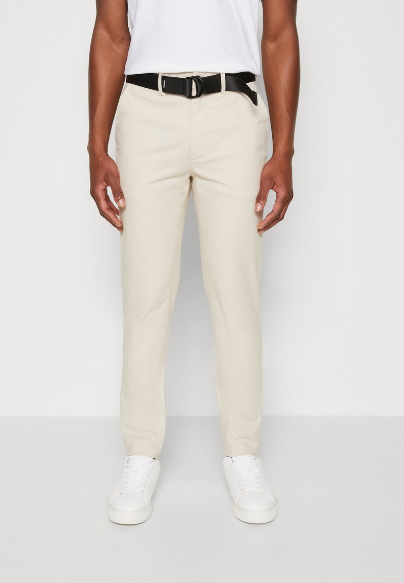 Pantaloni slim-fit beige chiaro in cotone, con una cintura nera dotata di una fibbia rotonda. Abbinati a scarpe da ginnastica bianche, creano un look casual.