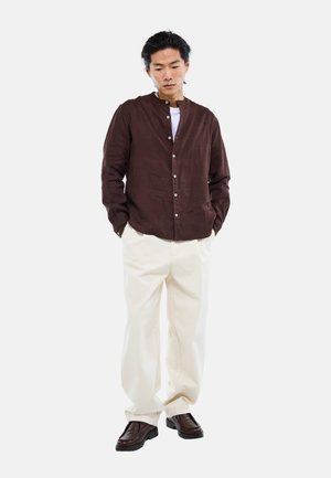 Un homme se tient les mains dans les poches, portant une chemise boutonnée marron foncé, un pantalon blanc à jambes larges, et des chaussures marron foncé sur un fond blanc uni.