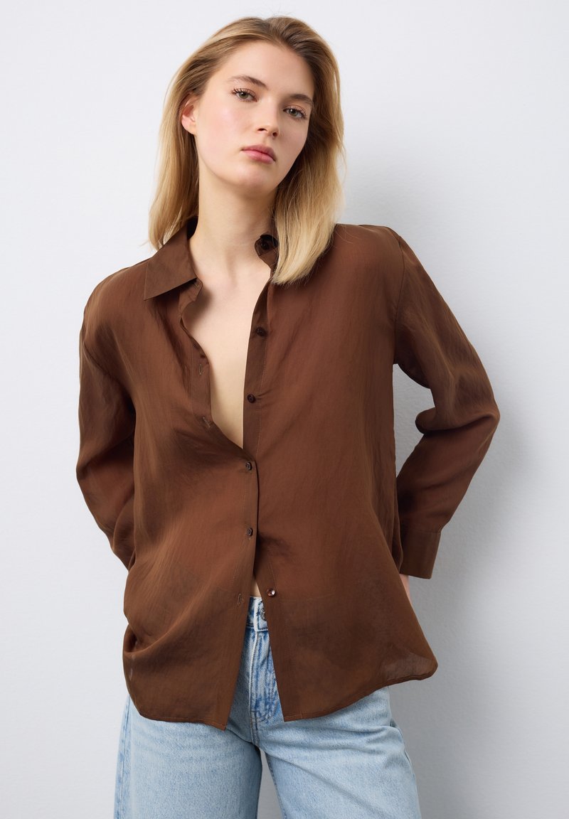 Junge Frau mit glattem blondem Haar, die ein lockeres braunes Hemd mit Knöpfen und hellblaue Jeans vor weißem Hintergrund trägt.