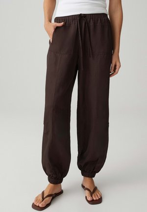 Donna che indossa pantaloni marrone scuro con coulisse, polsini elastici e tasche frontali, abbinati a infradito marroni e canotta bianca.