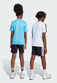 Dos niños están de espaldas, vistiendo camisetas deportivas de manga corta: una azul clara y otra blanca, con pantalones cortos negros y calcetines a rayas.