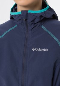Granatowa kurtka outdoorowa z kapturem, z kontrastowymi turkusowymi wykończeniami i zamkiem. Logo Columbia w srebrnym kolorze na lewej stronie klatki piersiowej.