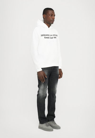 White hoodie with black text, paired with dark denim jeans. The jeans exhibit a faded finish. The model wears gray sneakers.