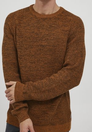 Homme portant un pull en maille marron texturé à manches longues, debout avec un bras croisé tenant le poignet opposé.