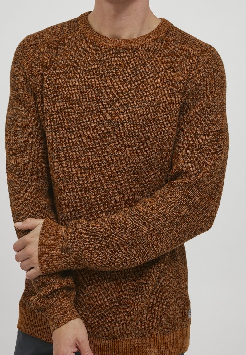 Homme portant un pull en maille marron texturé à manches longues, debout avec un bras croisé tenant le poignet opposé.