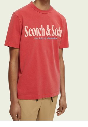 Mand iført en rød Scotch & Soda T-shirt med teksten "Free Spirit of Amsterdam" sammen med beige bukser mod en ensfarvet baggrund.