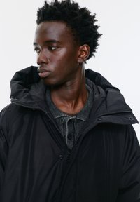 Tânăr cu păr scurt și creț, purtând o geacă neagră din puf și un pulover tricotat gri cu nasturi, privind spre stânga pe un fundal alb simplu.