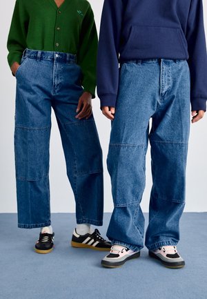 To personer iført løstsiddende, midtblå denimjeans med patchworkdetaljer. Den ene bærer en grøn cardigan, mens den anden har en marineblå hættetrøje på.