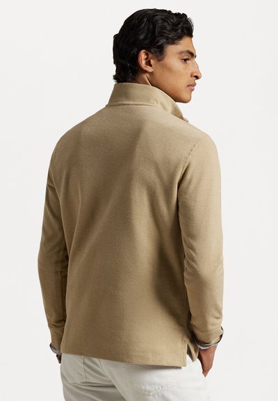 Tan long-sleeve polo shirt with a high collar, textured fabric, side slits for ease, and a slightly relaxed fit.