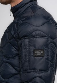 Marineblaue, gesteppten Pufferjacke mit hohem Kragen, ausgestattet mit strukturiertem Schulterbereich, gesticktem Muster und einem schwarzen Etikett am Ärmel.