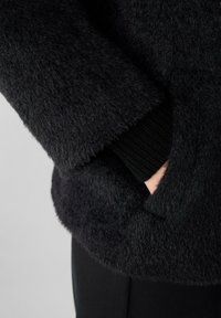 Černá, huňatá bunda se strukturovanou tkaninou, žebrovanými manžetami a kapsami, která zdůrazňuje jemný vzhled a elegantní design.