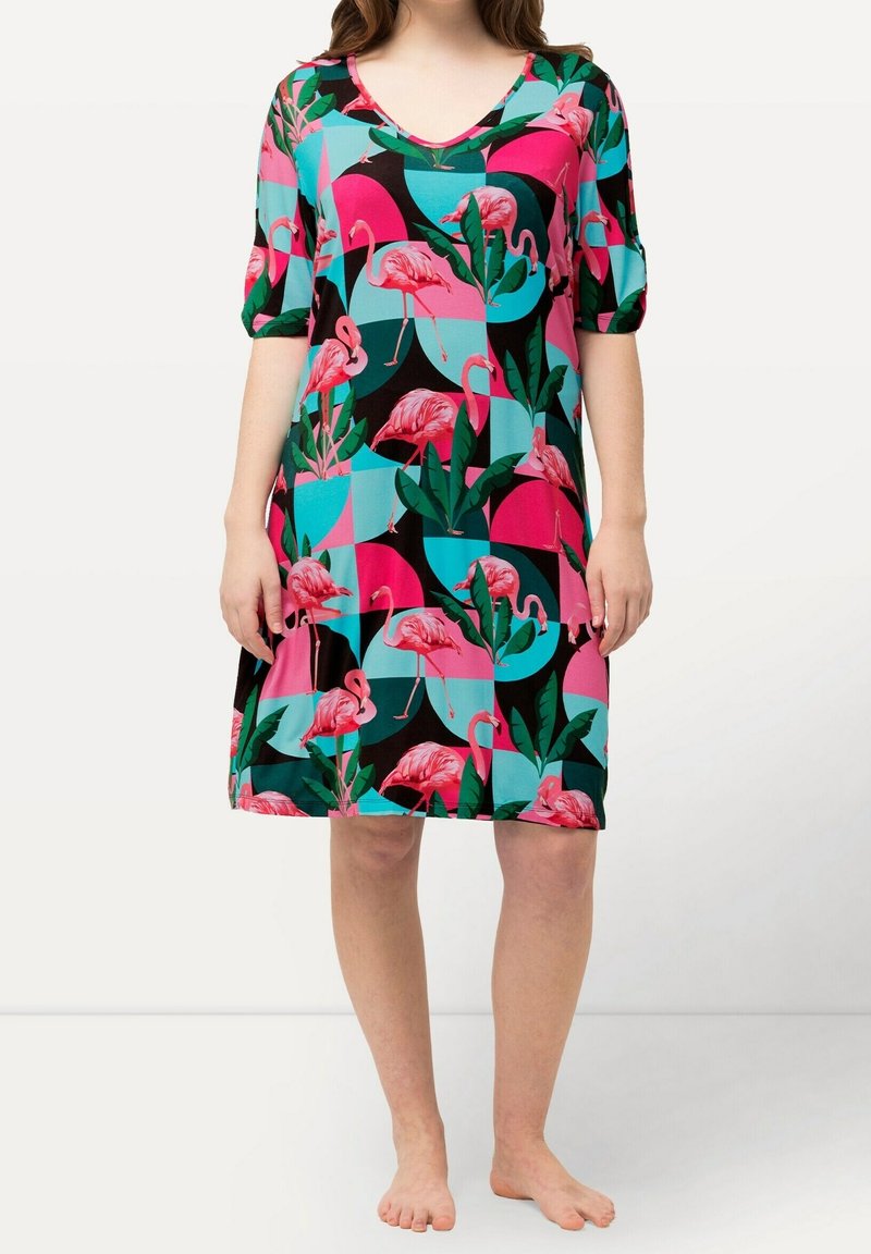 Femme portant une robe mi-longue avec un imprimé flamant rose et feuilles vertes sur un fond géométrique noir, rose et turquoise.