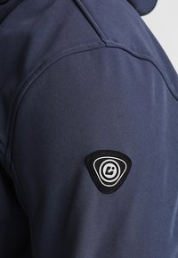 Épaule de veste bleu foncé avec un patch logo circulaire noir et blanc cousu près de la couture du haut du bras.