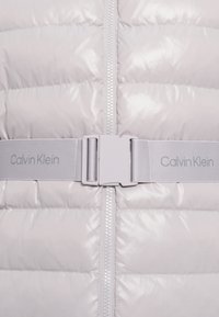 Svetlo šedá vypchávaná bunda s lesklou prešívanou textúrou, centrálnym zipsom a zodpovedajúcim pásom so zdobeným logom "Calvin Klein".