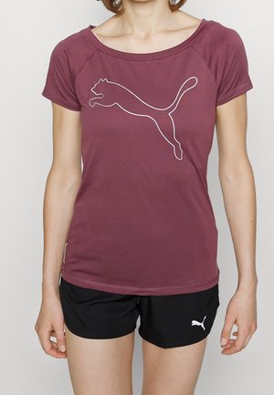 Femme portant un t-shirt Puma bordeaux avec logo puma bondissant et un short Puma noir avec logo puma blanc sur fond uni.