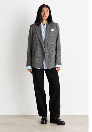 Blazer croisé à carreaux gris avec revers en cranté, associé à une chemise bleu clair boutonnée et à un pantalon noir à jambes larges. Des mocassins noirs complètent le look.