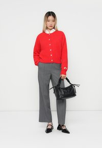 Cardigan rouge en tricot avec boutons, chemise blanche à col, pantalon gris à rayures fines, sac noir et chaussures noires à talons échancrés.