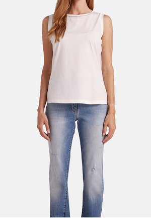 Femme portant un top blanc sans manches associé à un jean bleu clair délavé avec une légère usure sur un fond uni.