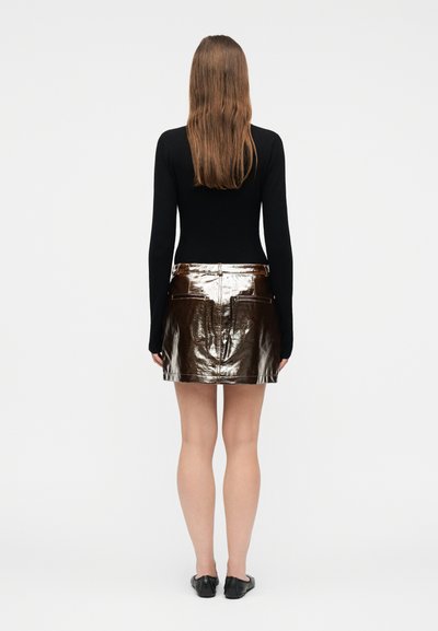 Femme aux longs cheveux bruns portant un haut noir à manches longues, une mini-jupe métallique marron et des chaussures plates noires, debout de dos sur un fond blanc.