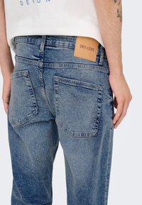 Jeans in denim blu con due tasche posteriori e una etichetta in pelle marrone chiaro sulla cintura con la scritta "ONLY & SONS" in rilievo.