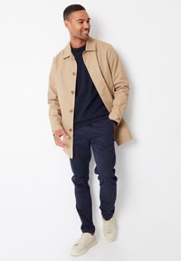 Beige lichtgewicht jack met bruine knopen, gedragen over een marineblauwe trui en marineblauwe broek, gecombineerd met crèmekleurige sneakers. Glad textuur en getailleerde pasvorm.