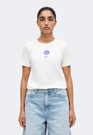 Femme aux cheveux foncés attachés porte un T-shirt blanc à manches courtes avec les logos bleus Paris et Nike, associé à un jean taille haute bleu clair.