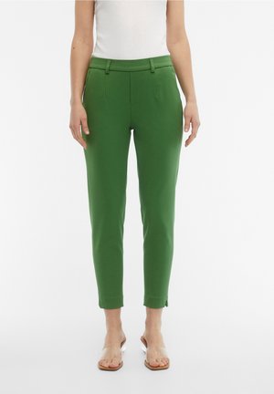 Woman wearing high-waist green slim-fit ankle pants with belt loops and front pockets, paired with clear strap sandals on a white background.