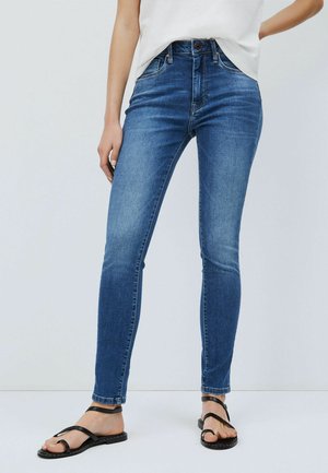 Kvinde iført tætsiddende blå jeans, hvid indstukket top og sorte flade sandaler med remme, stående foran en ensfarvet lys baggrund.