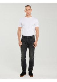 Mężczyzna stojący, ubrany w białą koszulę z krótkim rękawem, ciemnoszare dżinsy i czarne tenisówki z brązowymi podeszwami na jednolitym tle.