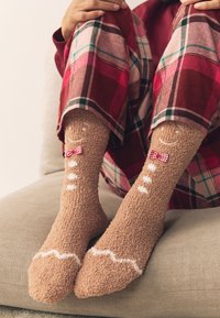 Vezelige, bruine sokken met thema van peperkoek, met een glimlachend gezicht, een rood geruit strikje en witte glazuurdetails, gedragen bij geruite broek.