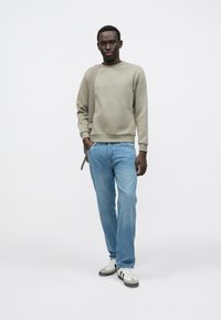 Felpa grigio chiaro con scollo rotondo, abbinata a jeans azzurri. Il modello indossa sneakers bianche e grigie e tiene una tracolla marrone.